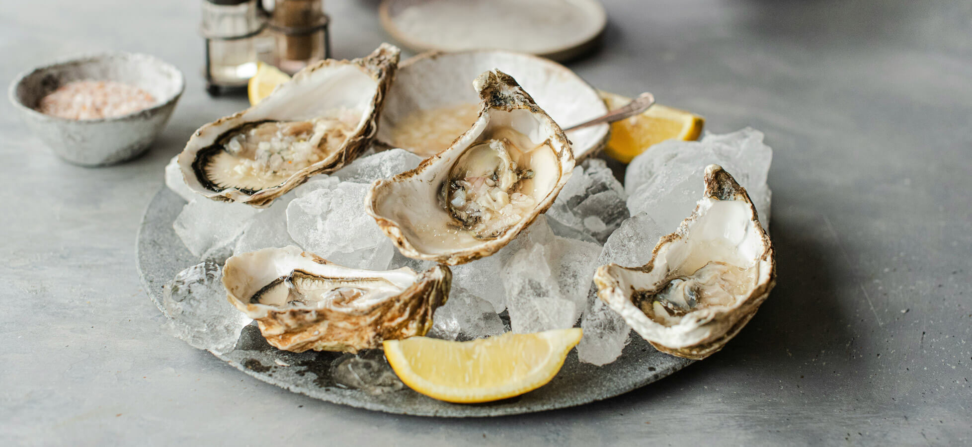Ein Teller mit frischen Austern auf Eis, umgeben von Zitronenscheiben. Die Austern sind geöffnet und präsentieren das schimmernde Fleisch. Im Hintergrund sind Gewürze in kleinen Schalen zu sehen. Die Gesamtpräsentation wirkt elegant und einladend.
