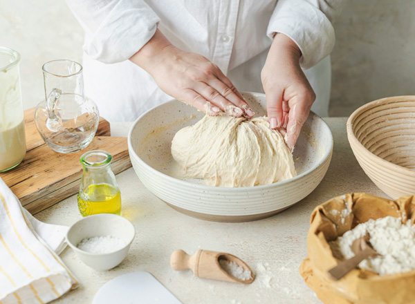 Eine Person knetet Teig in einer großen, flachen Schüssel. Auf dem Tisch sind verschiedene Zutaten wie Wasser, Öl und Mehl sichtbar. Die Hände sind leicht mit Teig bedeckt, während der Prozess des Brotbackens in Gang ist.