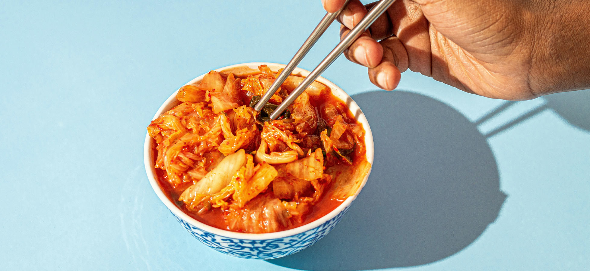 Eine Hand hält Essstäbchen und greift nach einer Portion Kimchi in einer Schüssel. Die Schüssel ist bunt dekoriert und steht auf einem hellblauen Tisch. Das Kimchi ist rot und enthält verschiedene Gemüsestücke.