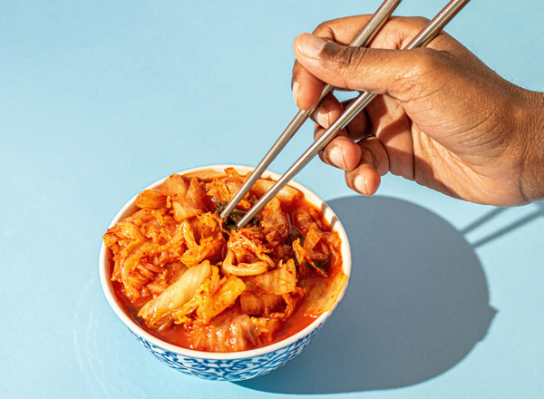Eine Hand hält Stäbchen, die ein Schälchen mit Kimchi greifen. Das Schälchen ist bunt verziert und der Hintergrund ist hellblau. Kimchi besteht aus eingelegtem und fermentiertem Gemüse, vorwiegend Chinakohl, und hat eine leuchtend rote Farbe.