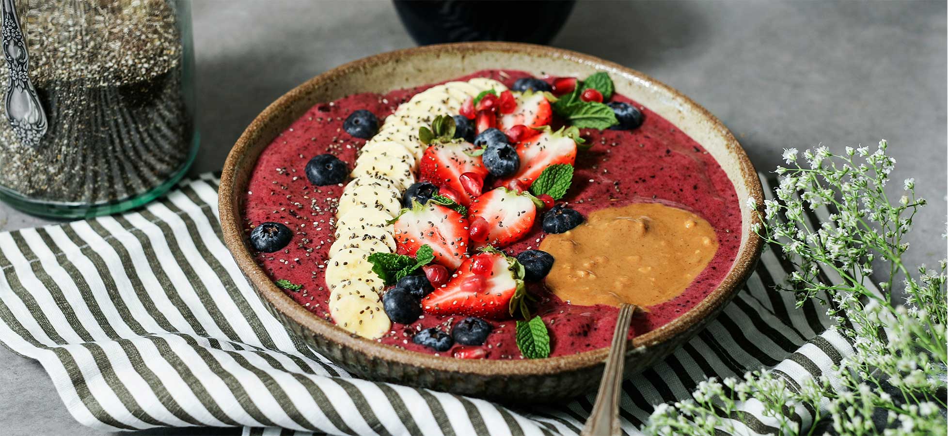 Eine Smoothie-Schüssel, garniert mit Bananenscheiben, Erdbeeren, Blaubeeren, Chiasamen, Minzblättern und Erdnussbutter.