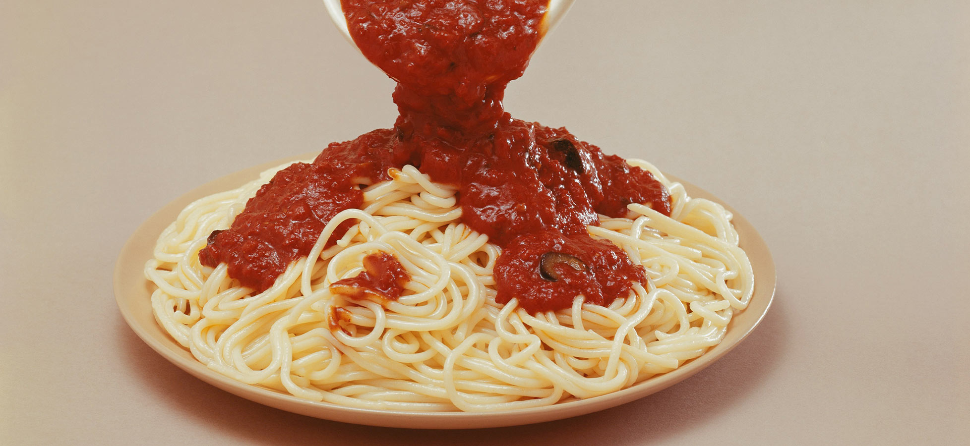 Ein Teller mit Spaghetti wird mit roter Tomatensoße übergossen. Die Spaghetti sind locker angeordnet und die Soße fließt gleichmäßig darüber. Der Hintergrund ist einfarbig, was den Fokus auf das Gericht legt.