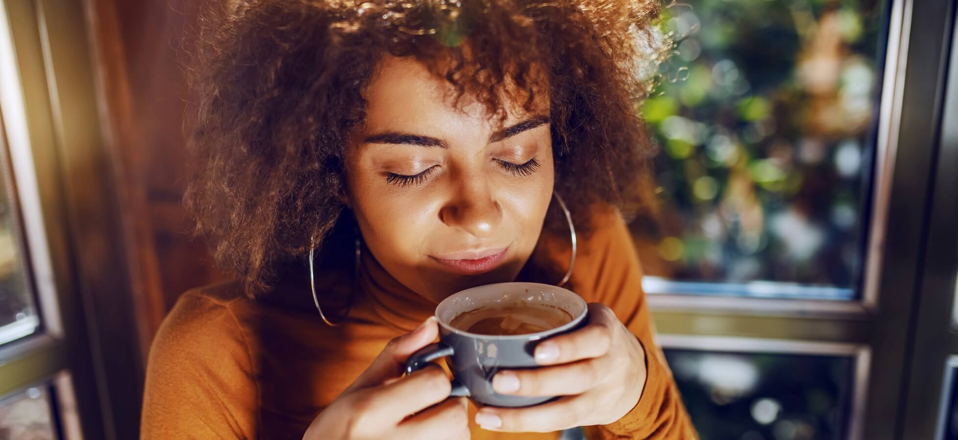 Eine junge Frau mit lockigem Haar sitzt am Fenster und hält eine Tasse in den Händen. Sie schließt die Augen und scheint den Duft des Getränks zu genießen. Die Umgebung ist hell erleuchtet und vermittelt eine warme, gemütliche Atmosphäre.