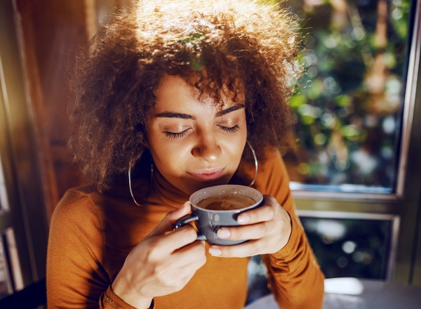 Eine junge Frau mit lockigem Haar sitzt an einem Tisch und hält eine Tasse in den Händen. Sie schließt die Augen und genießt den Duft des Getränks. Sonnenlicht strömt durch ein Fenster, das die Szene warm und einladend erscheinen lässt.