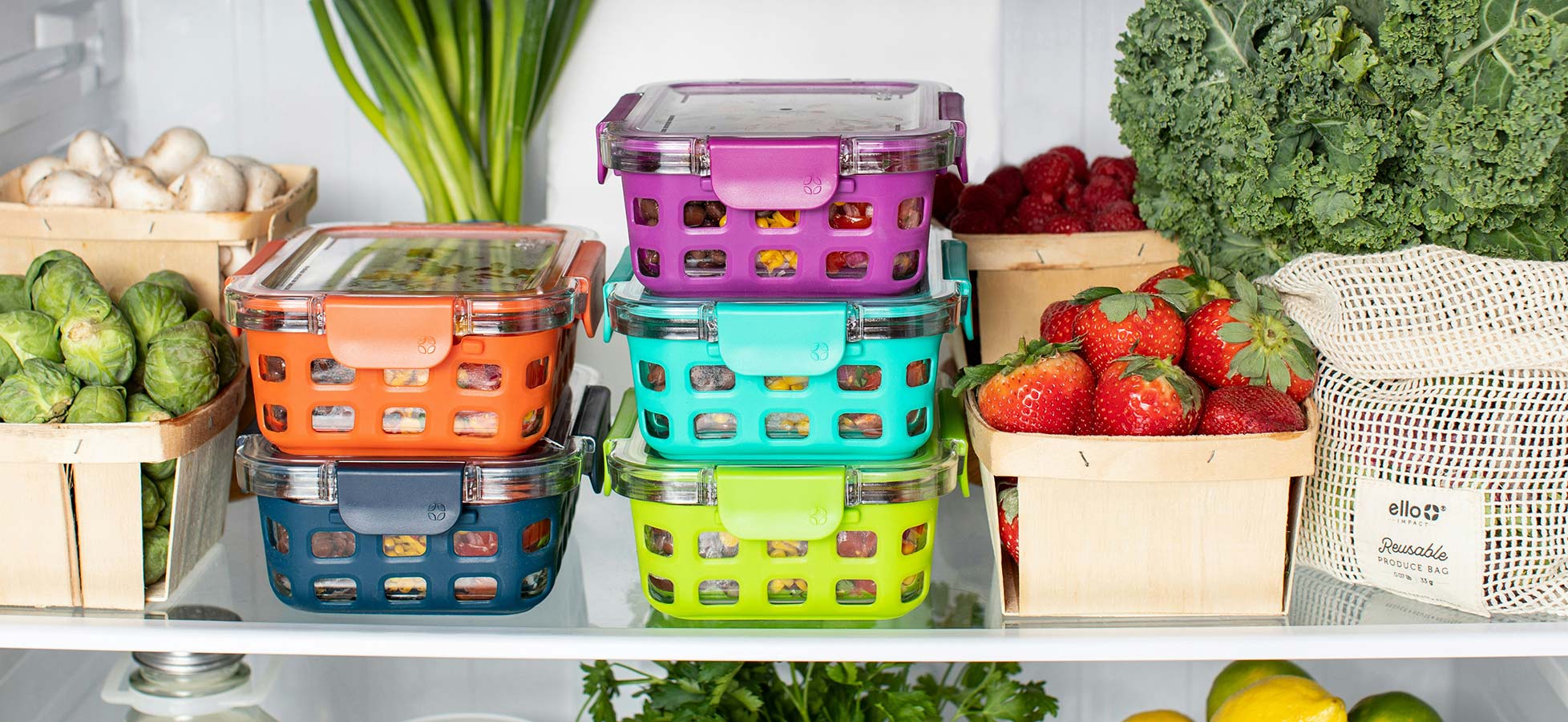 Im Kühlschrank sind mehrere Stapel von bunten, transparenten Aufbewahrungsboxen zu sehen. Diese enthalten frisches Gemüse und Obst wie Erdbeeren, Brokkoli und Rosenkohl. Einige kleine Holzkörbe sind ebenfalls mit frischen Lebensmitteln gefüllt.
