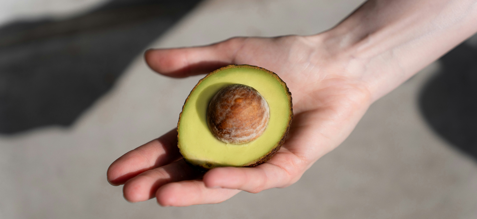 Eine Hand hält eine aufgeschnittene Avocado. Die fruchtige, grüne Innenseite ist gut sichtbar, während der runde, braune Kern in der Mitte liegt. Der Hintergrund ist neutral und unscharf.
