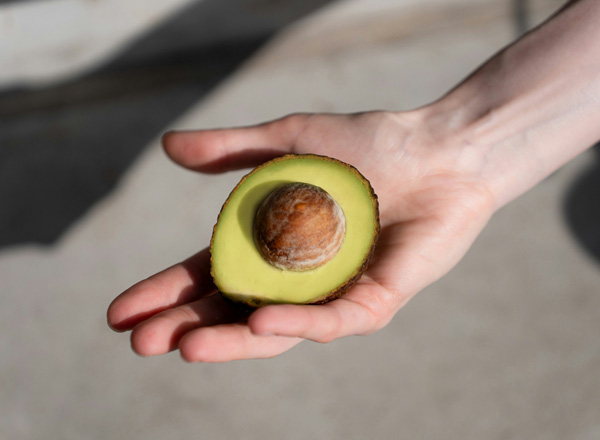Eine Hand hält eine halbierte Avocado, die ein braunes Kernstück zeigt. Das Fruchtfleisch ist leuchtend grün und die Schale ist dünn und dunkelgrün. Der Hintergrund ist unscharf und neutral.