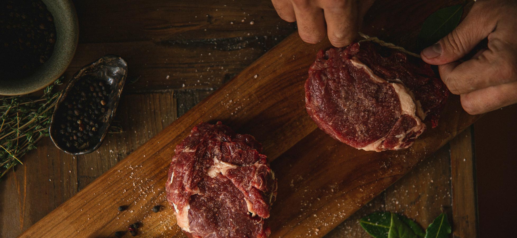 Auf einem Holzbrett liegen zwei Stücke rotes Fleisch, umgeben von grobem Salz, Pfefferkörnern und frischen Kräutern. Ein Olivenölbehälter und Knoblauch sind ebenfalls zu sehen, während eine Hand das Fleisch greift.