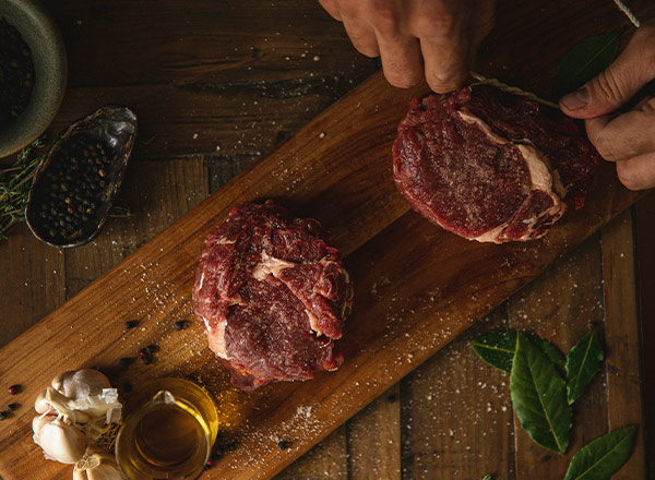Auf einem Holztisch liegen zwei Stücke rohes Fleisch. Eine Hand schneidet eines der Stücke. Neben dem Fleisch befinden sich Knoblauch, Öl und Gewürze. Die Szene vermittelt eine Vorbereitungsatmosphäre für das Kochen.