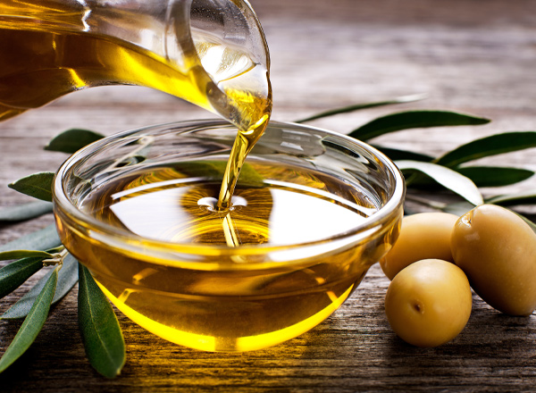 Ein Glas mit Olivenöl wird über eine Schale gegossen. Neben der Schale liegen grüne Oliven und einige Olivenblätter. Der Hintergrund zeigt eine hölzerne Oberfläche.