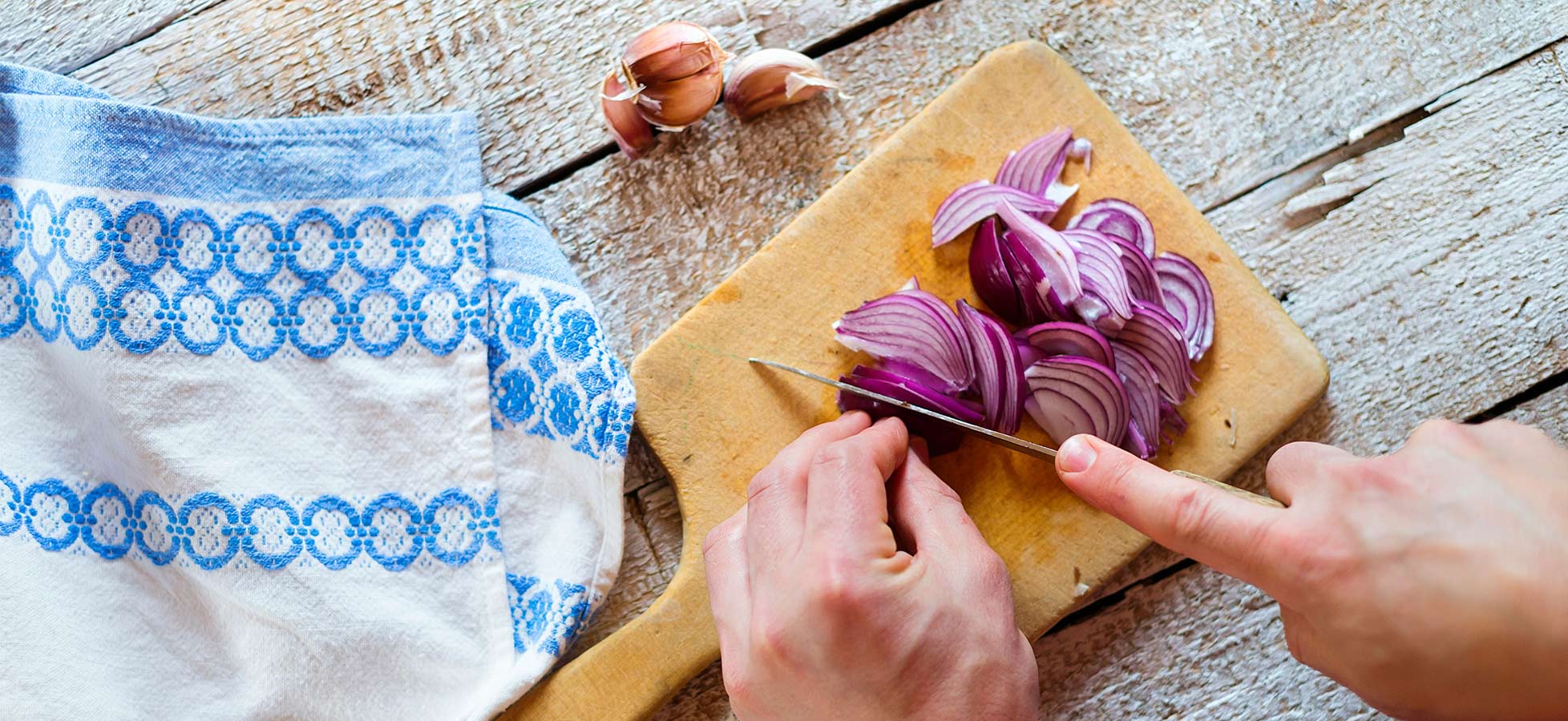 Eine Hand schneidet rote Zwiebeln auf einem Holzbrett, während eine Küchentuch mit blauem Muster daneben liegt. Im Hintergrund sind einige ungeschälte Knoblauchzehen sichtbar.