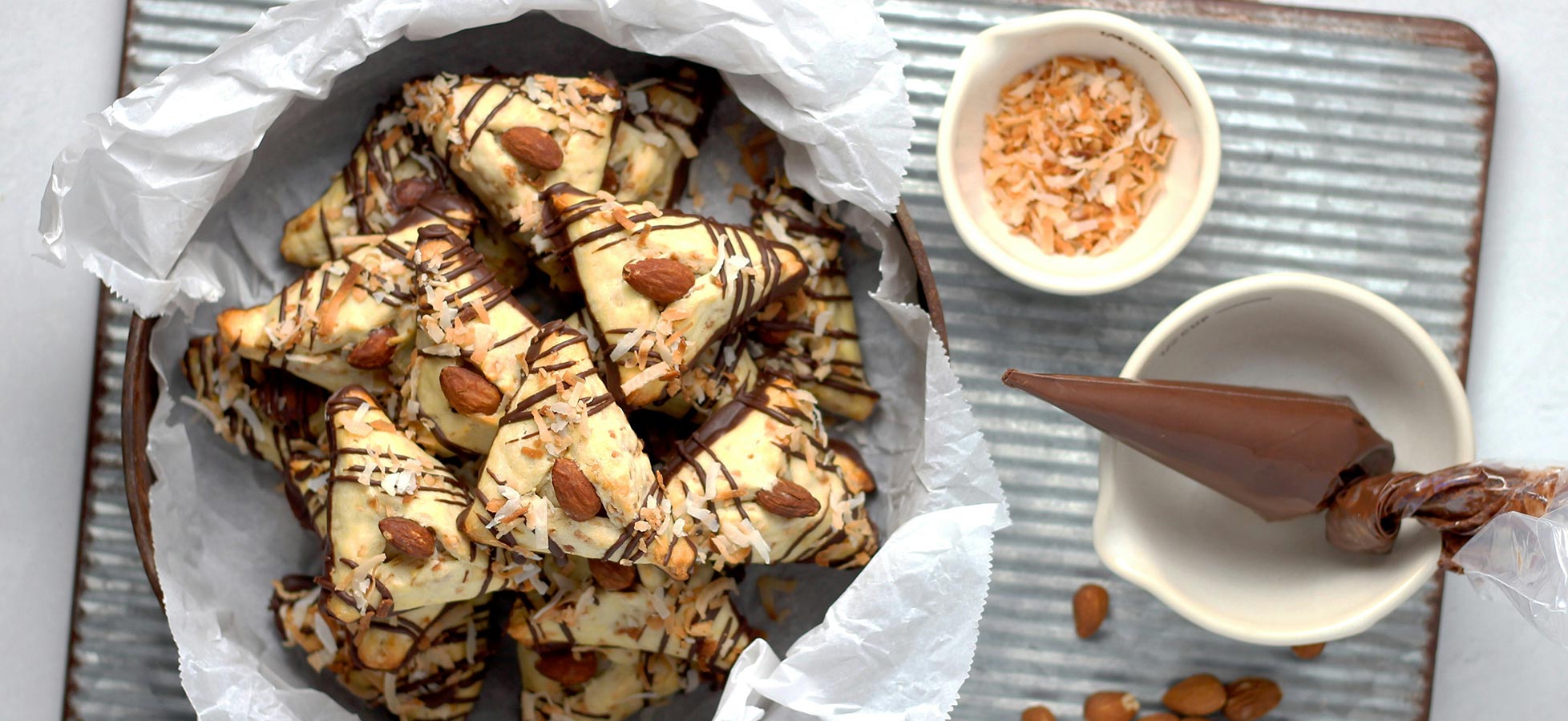 Ein Korb mit Hanfschnitten ist mit geschmolzener Schokolade und Mandeln dekoriert. Neben dem Korb befinden sich eine Schüssel mit Kokosraspeln und ein Spritzbeutel, der mit Schokolade gefüllt ist. Der Untergrund ist eine metallene Unterlage.
