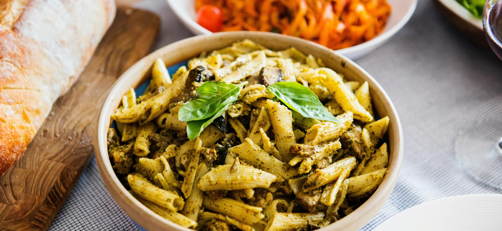 Eine Holzschüssel mit Penne-Pasta, bedeckt mit Pesto und garniert mit frischem Basilikum. Im Hintergrund sind verschiedene Gemüsegerichte und frisches Brot sichtbar. Die Szene vermittelt ein einladendes, rustikales Essensumfeld.