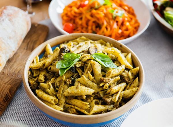 Eine Schüssel mit Penne-Pasta in Pesto-Sauce, garniert mit frischem Basilikum, steht im Vordergrund. Im Hintergrund sind andere Gerichte zu sehen, darunter eine rot gefärbte Pasta und ein Salat.