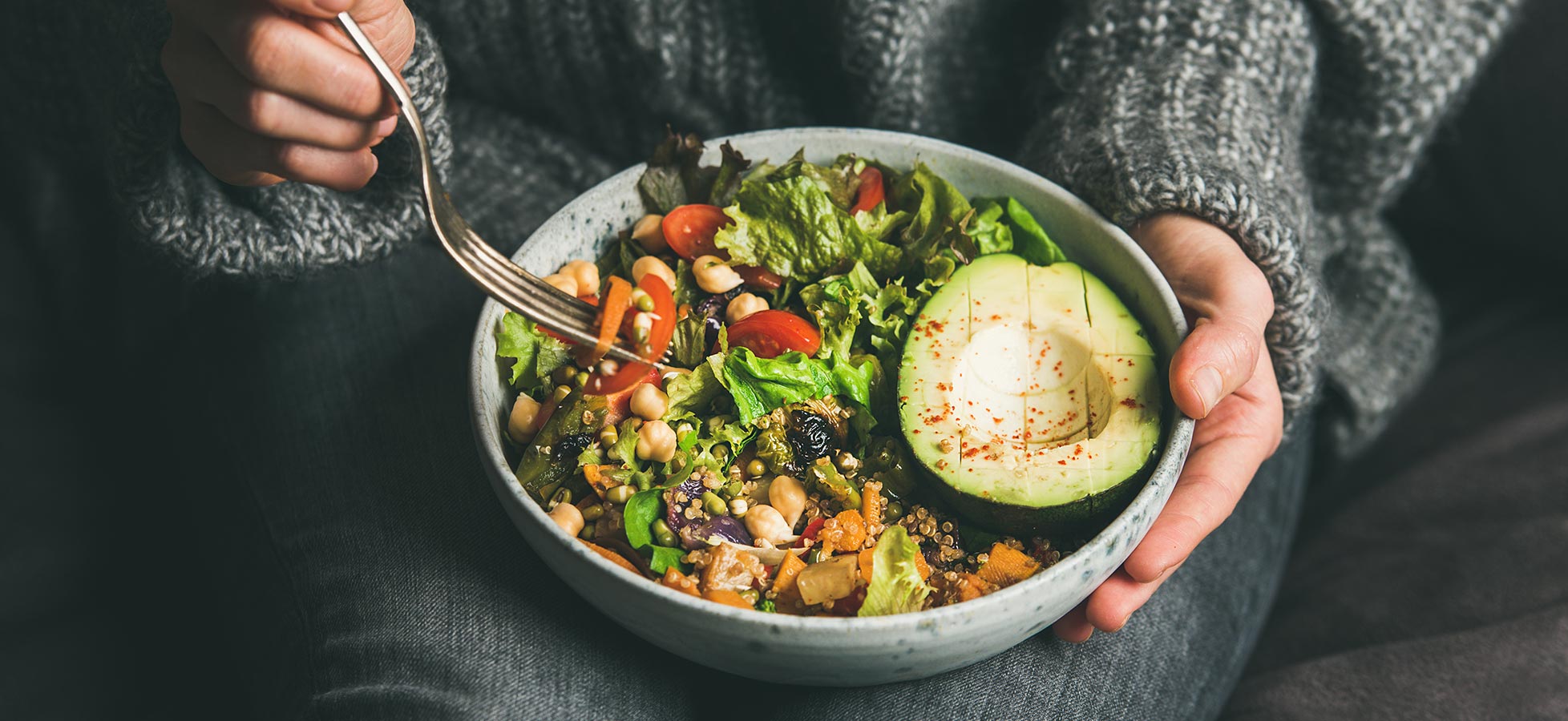 Eine Person hält eine Schüssel mit buntem Salat, der aus verschiedenen grünen Blattsalaten, Kirschtomaten, Karotten, Kichererbsen und einer halben Avocado besteht. Eine Gabel wird verwendet, um den Salat zu essen.