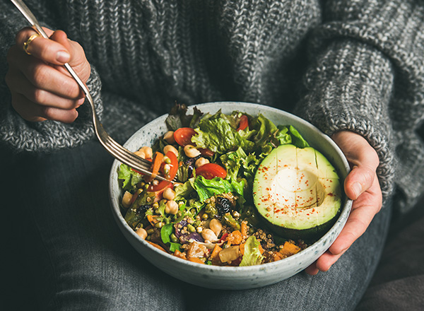 Eine Person hält eine Schüssel mit einem bunten Salat, der aus verschiedenen Blattsalaten, Kirschtomaten, Gurkenscheiben und Quinoa besteht. In der Schüssel sind auch Bohnen und Nüsse sichtbar. Die Person benutzt eine Gabel, um den Salat zu essen.