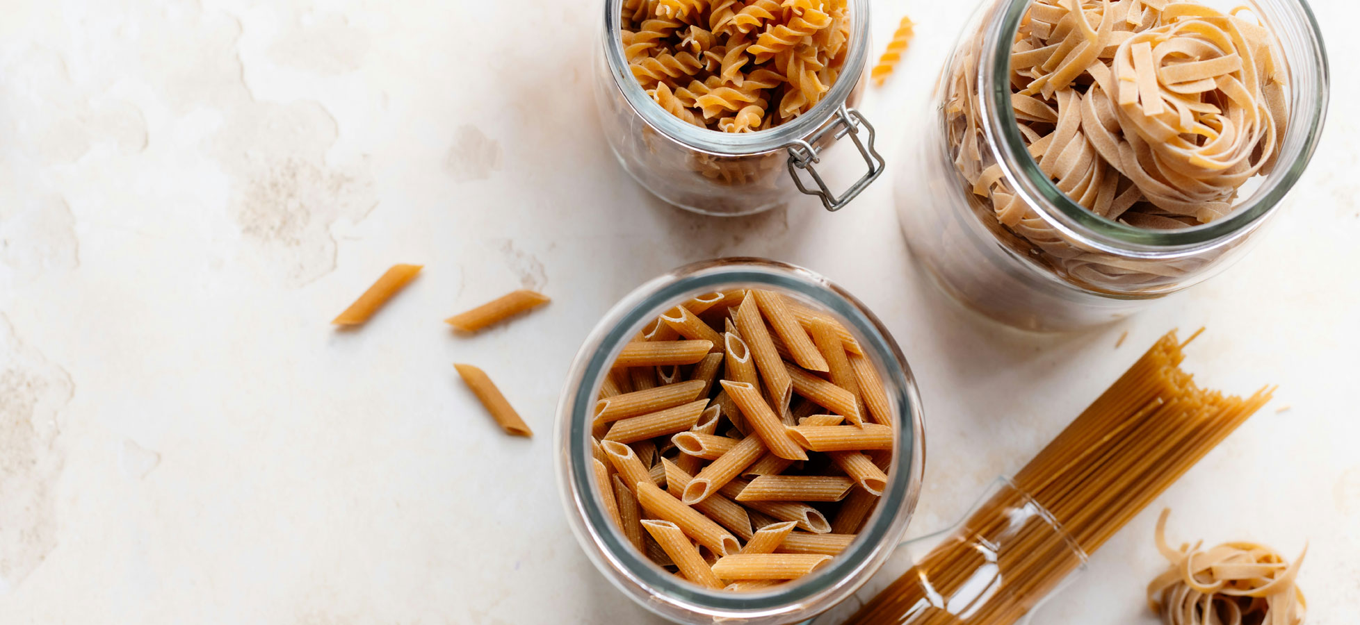 Drei Gläser mit verschiedenen Sorten Pasta stehen auf einer hellen Oberfläche. Eine Sorte ist röhrenförmig, die andere spiralförmig, und die dritte ist flach und bandförmig. Einige ungeordnete Pastastücke liegen auf der Oberfläche.