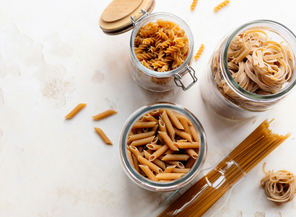 Drei Gläser mit verschiedenen Arten von ungekochter Pasta stehen auf einer neutralen Oberfläche. Im unteren Glas sind kurze röhrenförmige Nudeln, das mittlere Glas enthält spiralförmige Nudeln, und im oberen Glas sind breite Bandnudeln. Einige Nudeln liegen lose daneben.