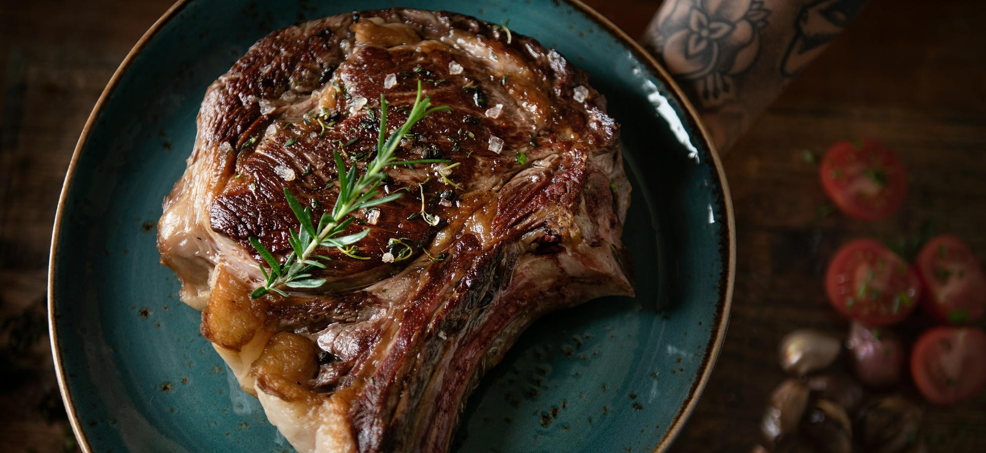 Ein saftiges Stück Steak auf einem blauen Teller, garniert mit frischen Kräutern und grobem Salz. Das Fleisch weist eine goldbraune Kruste auf und ist auf einem rustikalen Holzuntergrund platziert. Im Hintergrund sind einige frische Tomaten und Schalotten sichtbar.