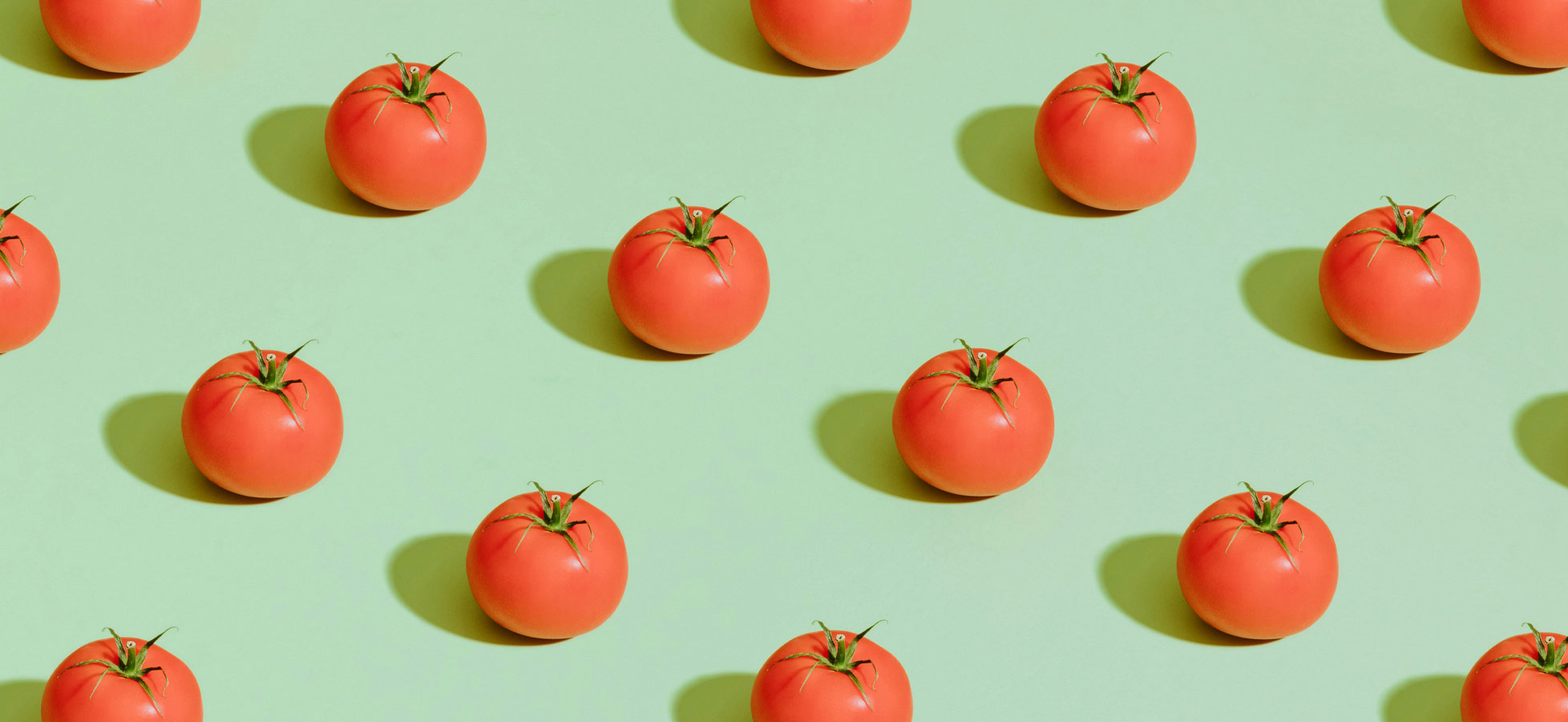 Eine Anordnung von roten Tomaten auf einer grünen Fläche. Die Tomaten sind glänzend und rund, jede mit grünen Blättern oben. Sie sind gleichmäßig verteilt und bilden ein harmonisches Muster.
