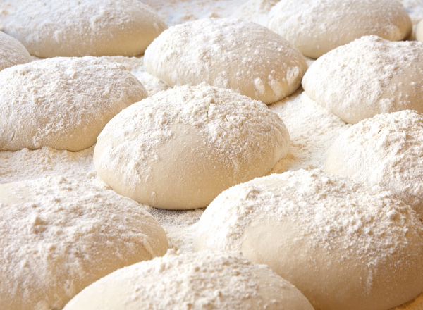 Mehrere rohe Teigstücke liegen auf einer bemehlten Oberfläche. Die Teigstücke sind rund geformt und teilweise mit Mehl bestäubt. Die Szene vermittelt den Eindruck, dass die Teigstücke zum Backen vorbereitet sind.