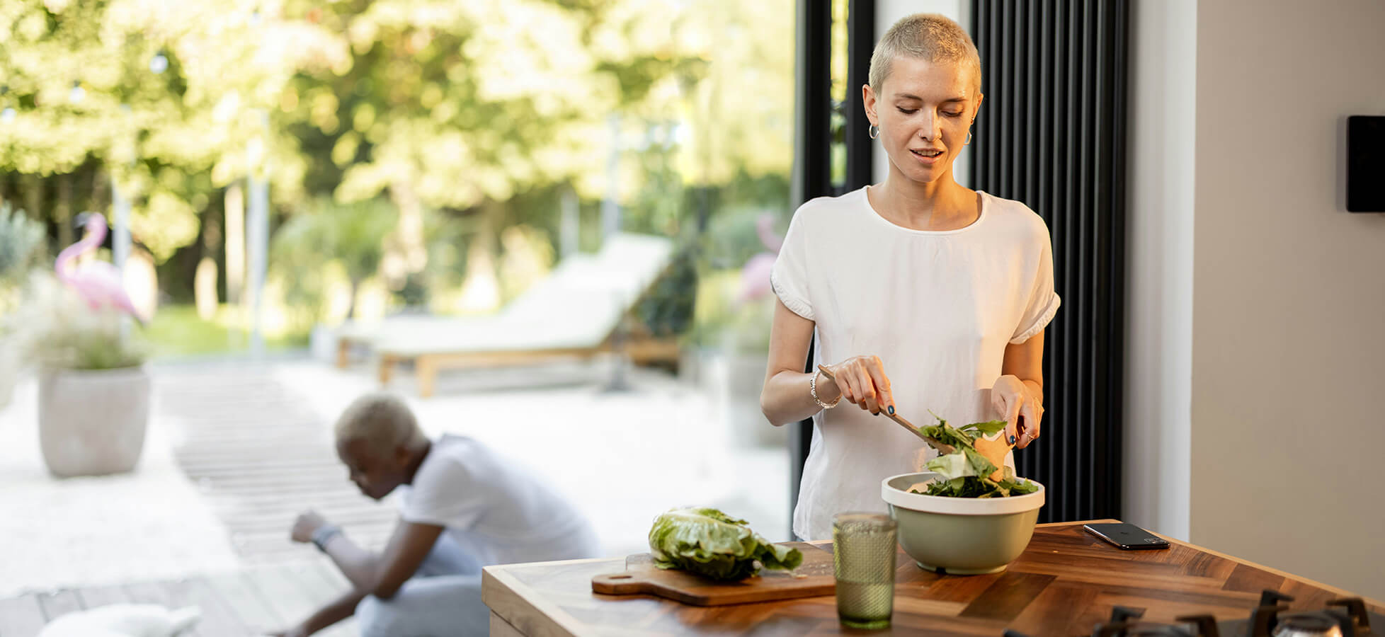Eine junge Frau bereitet in einer modernen Küche einen Salat in einer großen Schüssel vor. Sie trägt ein weißes T-Shirt und steht am Holztisch. Im Hintergrund ist eine Terrasse mit Gartenblick zu sehen.