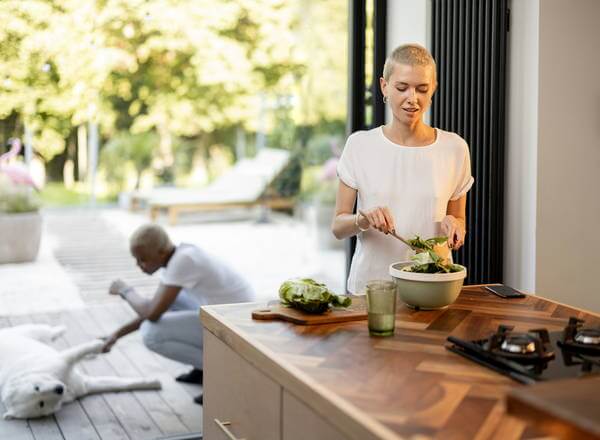 Eine Frau mit kurzem, blonden Haar bereitet einen Salat in einer modernen Küche vor, während im Hintergrund ein Mann auf der Terrasse mit einem Hund spielt. Der Raum ist hell und großzügig, mit Blick auf einen Garten.