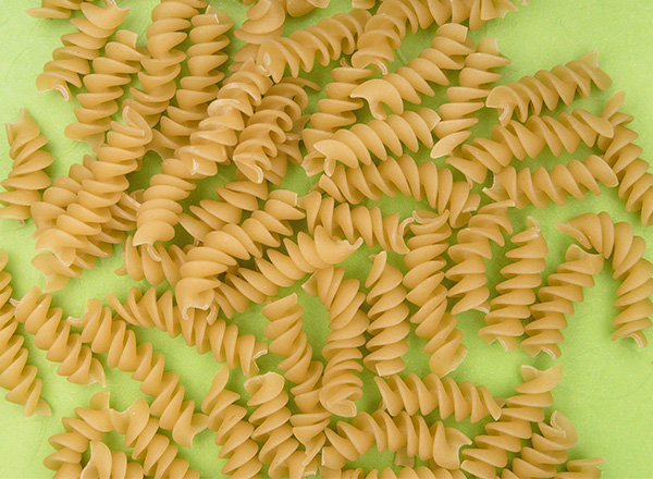 Eine große Menge von beige Farfalle-Nudeln liegt verstreut auf einer hellgrünen Fläche. Die Nudeln haben eine spiralenartige Form und sind in verschiedenen Positionen angeordnet.