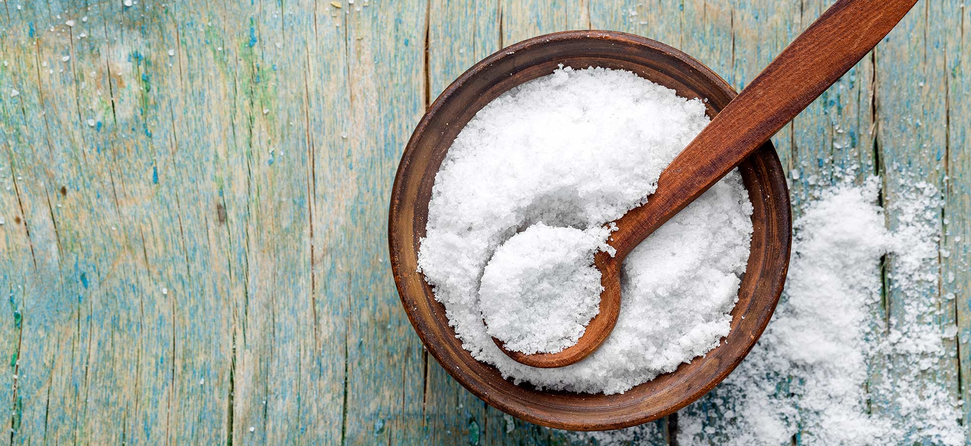 Eine Schale aus Holz enthält grobkörniges Salz, aus dem ein Holzlöffel herausragt. Einige Salzkrümel liegen auf der Oberfläche des Untergrundes, der in Holzoptik gestaltet ist.
