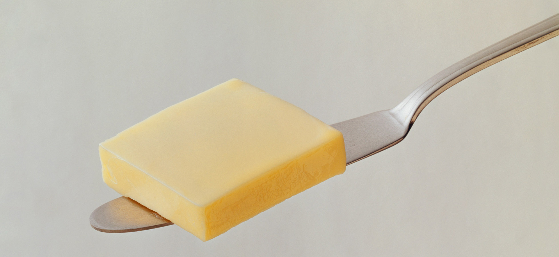 Ein Stück Butter liegt auf einem Edelstahl-Messer. Die Butter hat eine rechteckige Form und eine glatte, gelbliche Oberfläche. Der Hintergrund ist hell und unauffällig, was die Butter in den Vordergrund rückt.