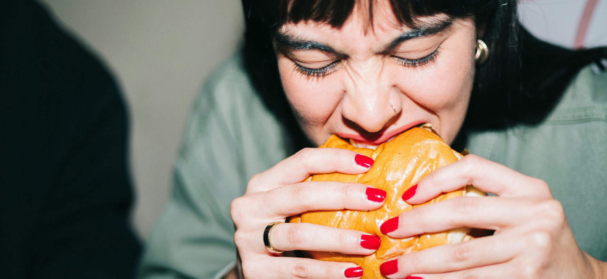 Eine Person mit dunklen Haaren und roten Nägeln hält einen großen Burger und bereitet sich darauf vor, hinein zu beißen. Sie sieht konzentriert und hungrig aus. Der Hintergrund ist unscharf und lenkt nicht von der Handlung ab.