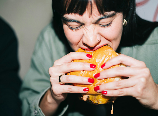 Eine Person mit kurzen, dunklen Haaren und rotem Nagellack beißt in einen großen Burger. Ihr Gesicht zeigt einen konzentrierten Ausdruck, während Käse von dem Burger tropft. Die Hände halten den Burger fest.