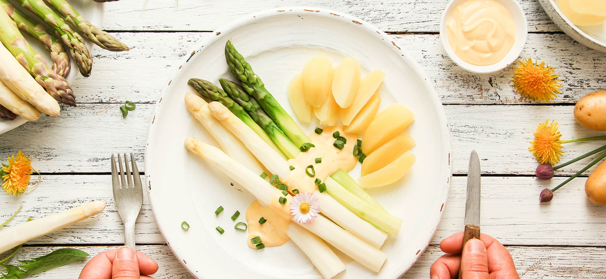 Ein weißer Teller mit gekochtem Spargel, grünem Spargel und Kartoffelscheiben, garniert mit Schnittlauch und einer Blüte. Zwei Hände halten Besteck, bereit zum Essen. Weitere Spargel und Kartoffeln liegen auf dem Tisch.