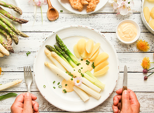 Ein Teller mit weißem und grünem Spargel, umgeben von gekochten Kartoffeln. Der Spargel ist mit grünen Zwiebelringen garniert und eine Blüte ziert das Gericht. Am Rand des Tellers liegt eine Schale Sauce und einige Hähnchenstücke sind sichtbar.