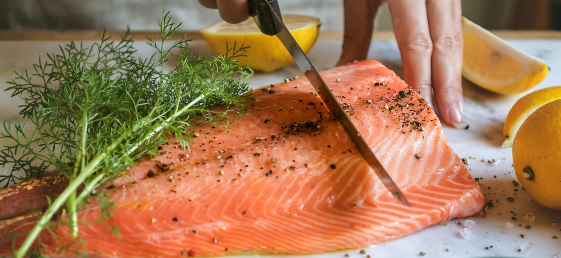 Ein Koch schneidet frischen Lachs auf einem Schneidebrett. Der Lachs ist mit Pfeffer und frischen Kräutern garniert, umgeben von Halben einer Zitrone. Die Szene vermittelt eine appetitliche Atmosphäre, die auf Zubereitung und Genuss von frischem Fisch hinweist.