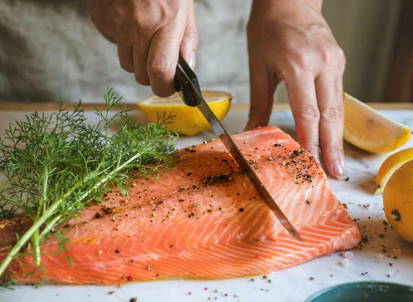 Eine Hand schneidet mit einem Küchenmesser ein Stück rohen Lachs auf einem Schneidebrett. Neben dem Lachs liegen frische Zitronenscheiben und einige Kräuter. Der Lachs ist mit Gewürzen bestreut, was auf die Zubereitung von Fischgerichten hinweist.