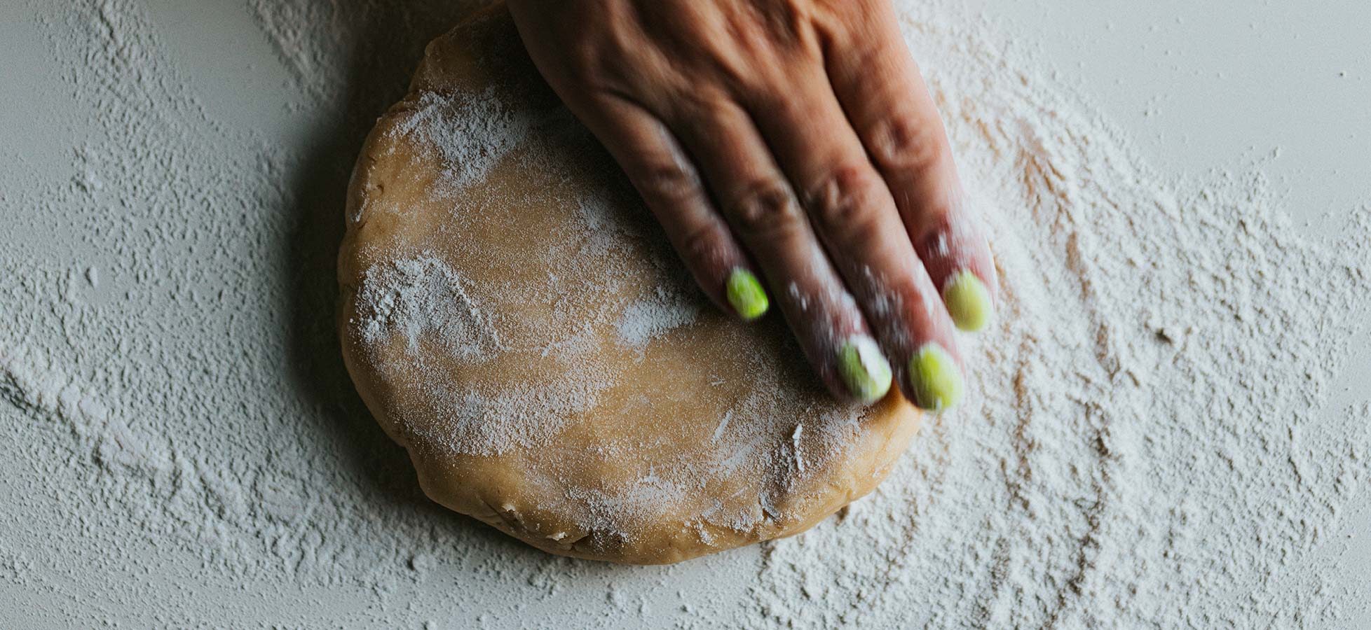 Eine Hand glattet einen runden Teigblock, der mit Mehl bestäubt ist. Der Hintergrund ist mit zusätzlichem Mehl bedeckt, was auf den Backprozess hinweist.