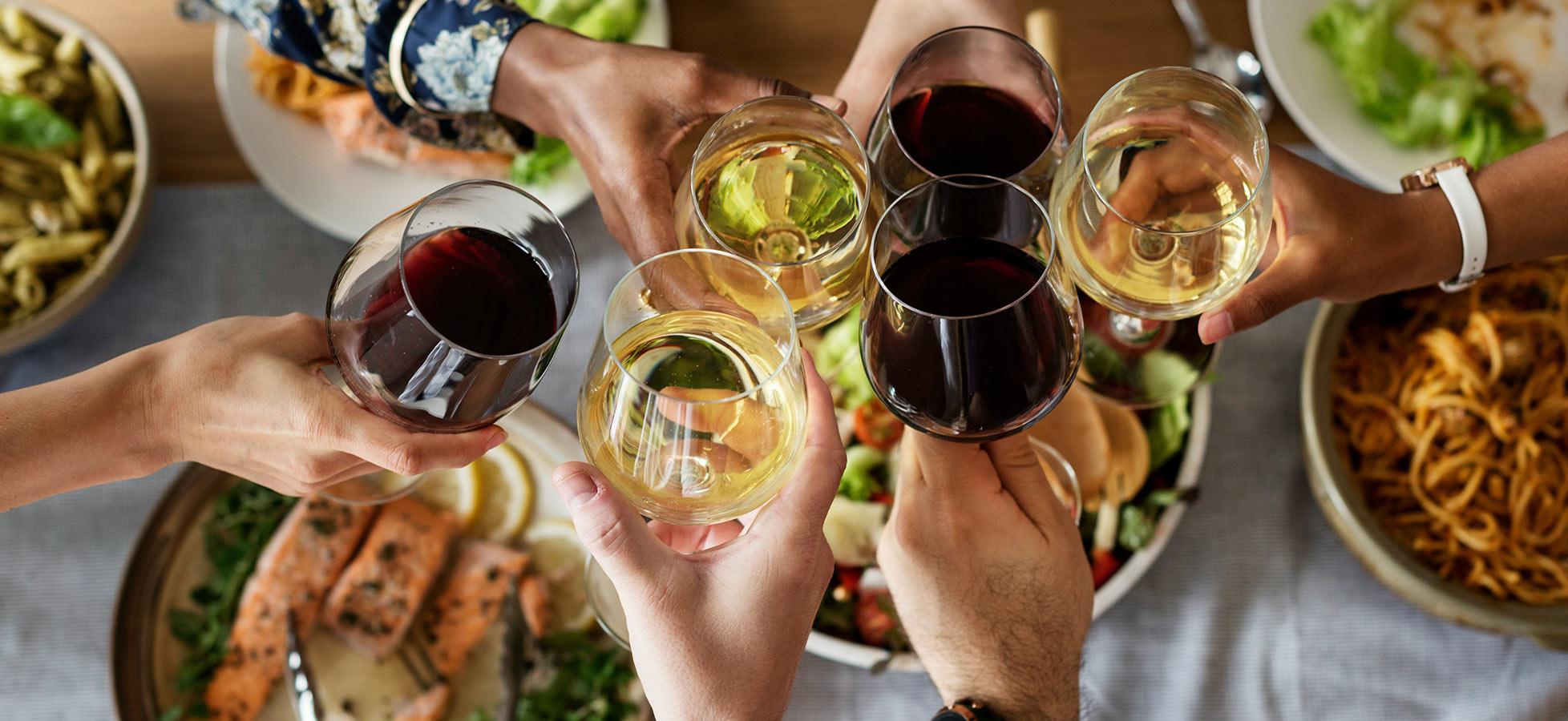 Sechs Personen stoßen mit Weingläsern an. Die Gläser enthalten roten und weißen Wein. Im Hintergrund sind Teller mit verschiedenen Speisen zu sehen, darunter Fisch und Salat. Die Szene vermittelt eine festliche und gesellige Atmosphäre.