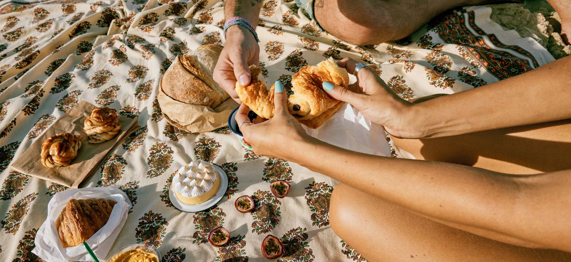 Zwei Menschen sitzen auf einem bunten Tuch und teilen frische Backwaren. In ihren Händen halten sie ein Croissant und ein Stück eines anderen Gebäckstücks. Verschiedene süße Leckereien und ein ungeschnittenes Brot sind ebenfalls auf dem Tuch verteilt.