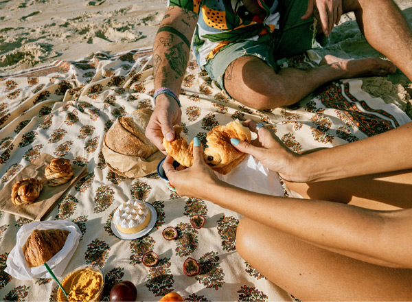 Zwei Personen sitzen auf einer Decke am Strand, während sie sich Gebäckstücke wie Croissants und Brötchen reichen. Vor ihnen sind verschiedene Snacks und eine kleine Torte zu sehen. Die Atmosphäre wirkt entspannt und gesellig.