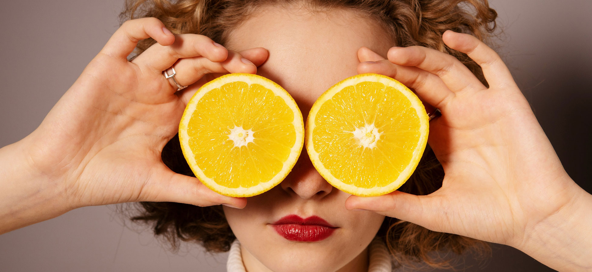 Eine Person hält zwei halbierte Zitronen vor ihr Gesicht. Die Zitronen verdecken die Augen, während der Rest des Gesichts sichtbar ist. Die Person hat lockiges Haar und trägt roten Lippenstift. Der Hintergrund ist neutral gehalten.
