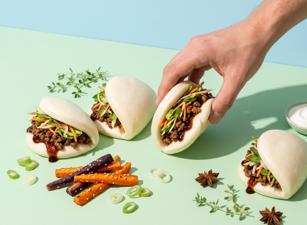 Eine Hand greift nach einem gefüllten Bao-Brötchen, das mit Gemüse und einer dunklen Sauce belegt ist. Im Vordergrund liegen bunte Karotten und Kräuter. Die Szene vermittelt ein frisches und appetitliches Essensangebot.