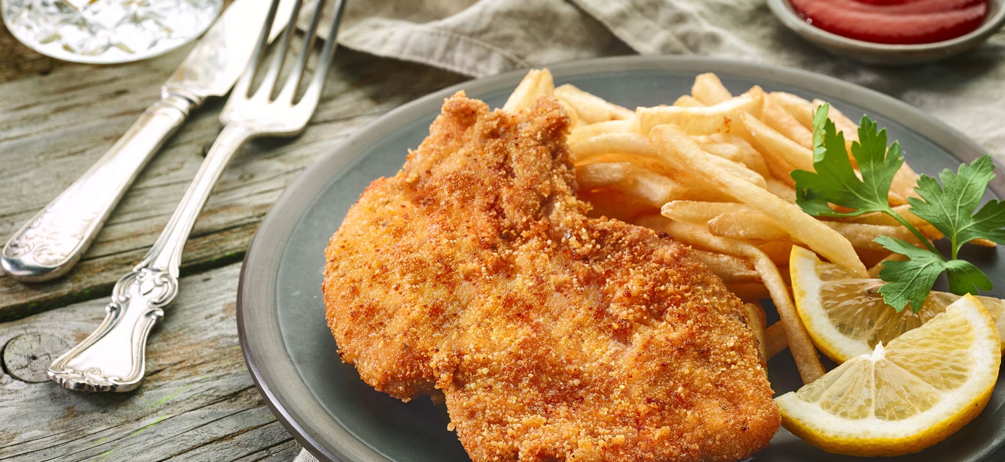 Ein Teller mit paniertem Schnitzel liegt neben einer Portion Pommes Frites. Auf dem Teller sind auch einige Zitronenscheiben und ein paar frische Kräuter zu sehen.