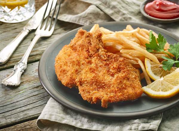Ein Teller mit paniertem Schnitzel liegt auf einem grauen Tuch. Daneben befinden sich Pommes frites, eine Zitronenscheibe und etwas frische Petersilie. Im Hintergrund sieht man Ketchup in einem kleinen Schälchen.