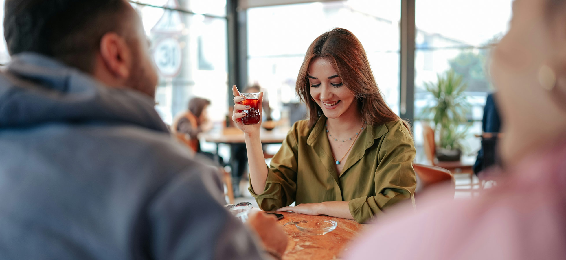 Eine lächelnde Frau sitzt an einem Tisch in einem Café und hält ein Glas mit einem Getränk in der Hand. Sie trägt eine grüne Bluse und schaut freundlich zu einer Person, die sich ihr gegenübersitzt. Der Raum ist hell und einladend, mit anderen Gästen im Hintergrund.