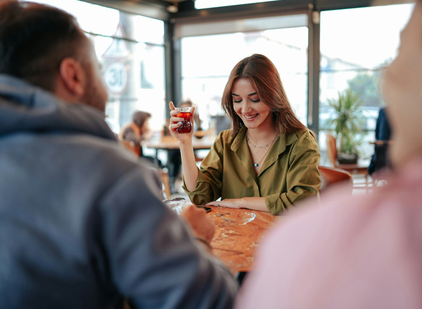 Eine lächelnde Frau in einem grünen Hemd hält ein Glas mit einem dunklen Getränk in der Hand. Sie sitzt an einem Tisch in einem Café und schaut auf das Getränk. Im Hintergrund sind unscharfe Silhouetten von weiteren Personen und Pflanzen sichtbar.