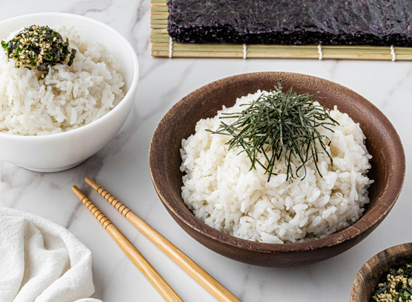 Eine Schüssel dampfender, lockerer Reis steht auf einem matschigen Untergrund. Neben der Schüssel liegen essbare Holzstäbchen. Der Fokus liegt auf dem heißen Reis, der appetitlich und frisch aussieht.