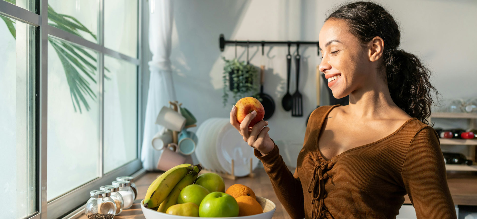 Eine lächelnde Frau hält einen Apfel in der Hand, während sie in eine Schüssel mit buntem Obst schaut. Im Hintergrund sind eine Fensterfront und Küchenutensilien sichtbar. Die Atmosphäre ist hell und einladend.