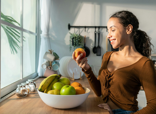 Eine Frau steht an einem Tisch und hält einen Apfel in der Hand. Vor ihr befindet sich eine große Schüssel mit verschiedenen Früchten, darunter Bananen, grüne und orange Äpfel. Sie lächelt, während sie den Apfel betrachtet, mit Sonnenlicht, das durch das Fenster strömt.