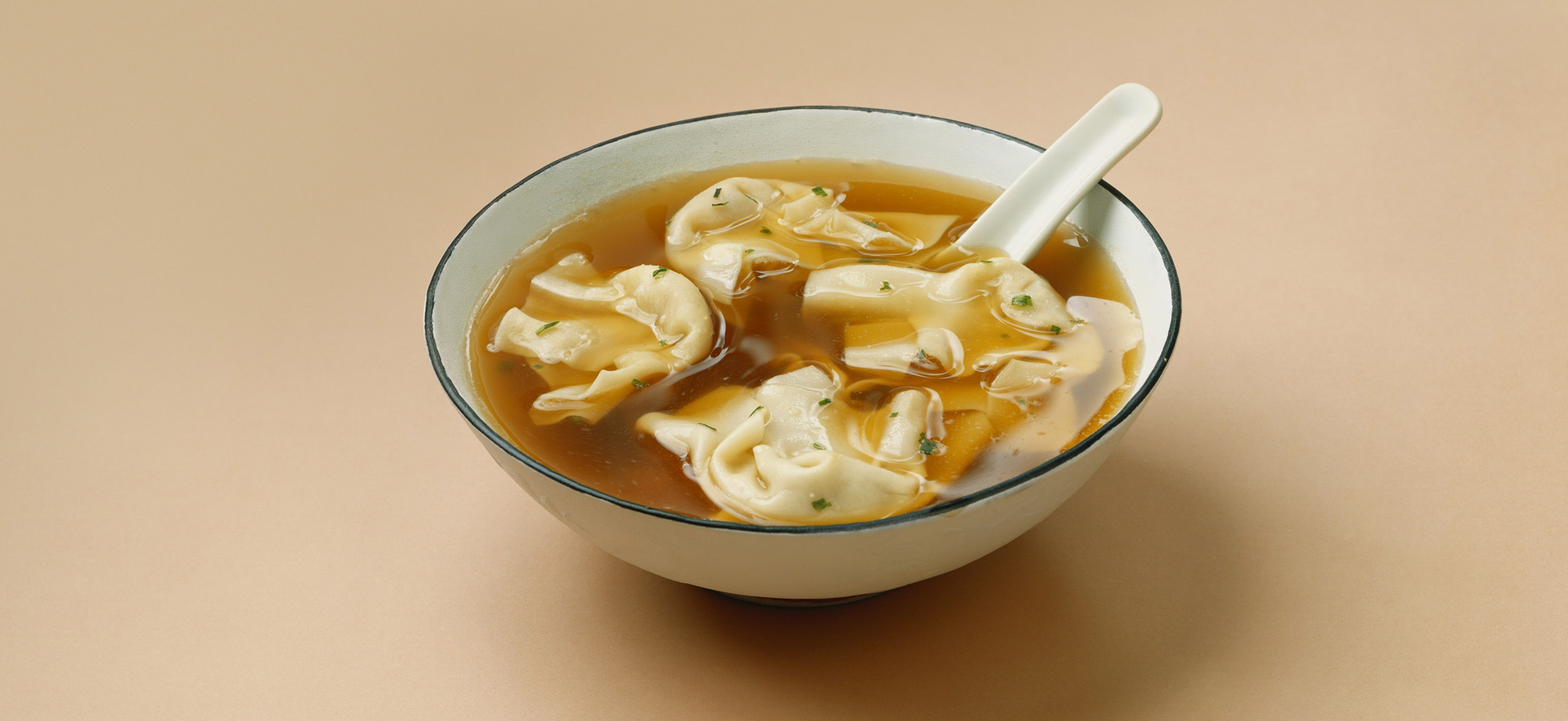 Eine Schüssel mit klarer Brühe, in der mehrere gefüllte Teigtaschen schwimmen. Die Brühe ist leicht goldfarben und wird von frischen Kräutern verziert. Ein weißer Löffel liegt in der Schüssel. Der Hintergrund ist ein sanfter Beigeton.